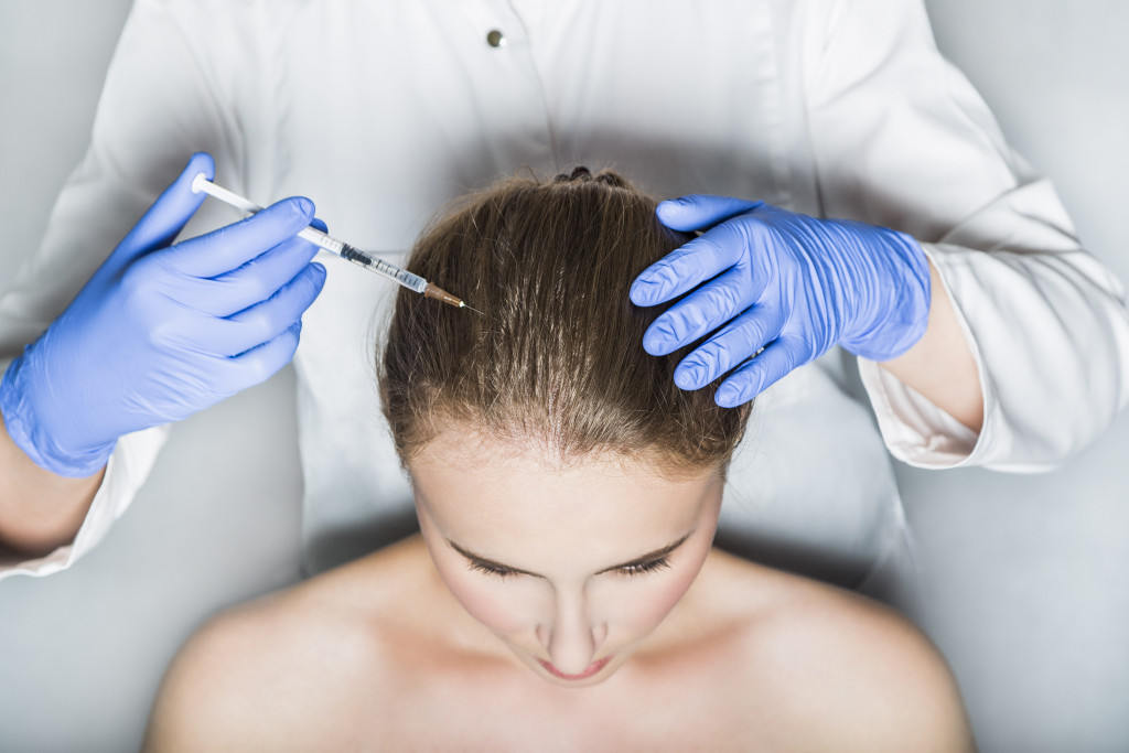 Doctor aesthetician with blue medical gloves and white medical gown makes hyaluronic acid rejuvenation beauty injections in the head of female patient for hair growth and to prevent boldness.