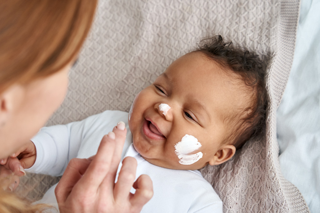mother putting sunscreen to baby's smiling face in the bed
